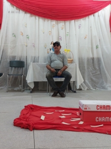 Foto de um senhor de óculos, motorista do ônibus escolar. Ao fundo uma cortila branca e vermelha, no centro da sala um pano vermelho com papeis e uma caixa vermelha e branca